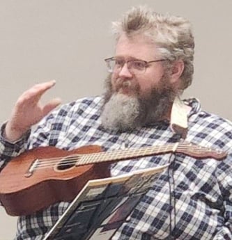 Picture of Caleb, the teaching artist. He  is wearing a ukulele strapped around his neck, and has him arm raised. He has light hair, a beard, and glasses.