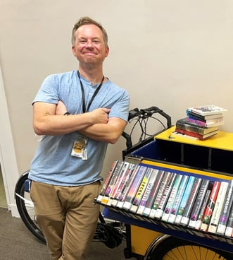 Picture of Kevin, the library worker. He has short blonde hair and has his arms crossed while leaning against a cart of books.