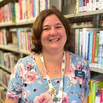 Headshot of library worker, Sarah. She has a light brown bob and is wearing a floral shirt. She is standing in front of a bookshelf.