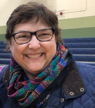 Picture of Gwen, the teaching artist, smiling. She has short brown hair and dark-framed glasses. She is wearing a blue, green, and purple scarf.