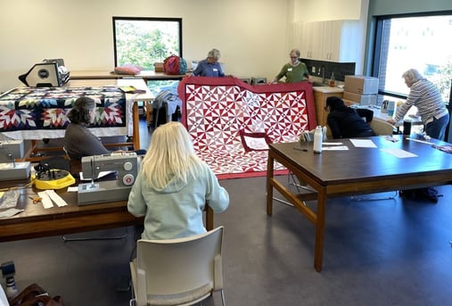 Two older adults hold up a large quilting tapestry in front of other adults sitting at tables.