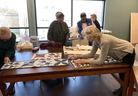 A group of older adults lean on tables while looking at quilting tapestries. 