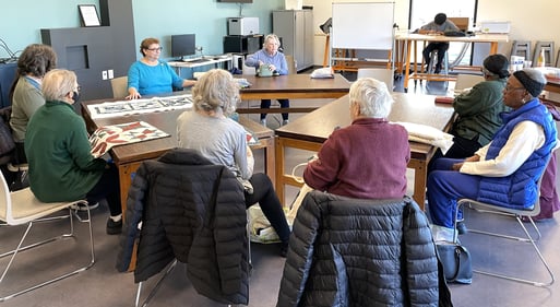 A group of older adults sit in chairs around tables while listening to instructor, Gwen. Quilting fabric is laying on the tables.
