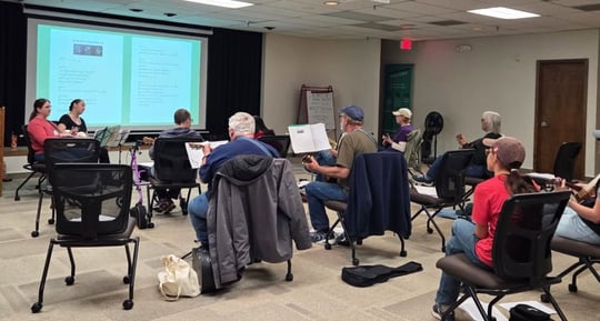 A group of older adults practice ukulele while listening to two instructors.