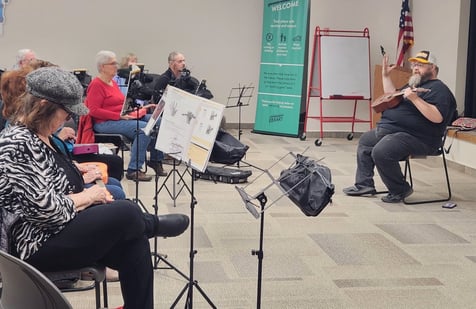 Teacher, Caleb Smith sits in a chair holding a Ukulele in front of a group of older adults listening.