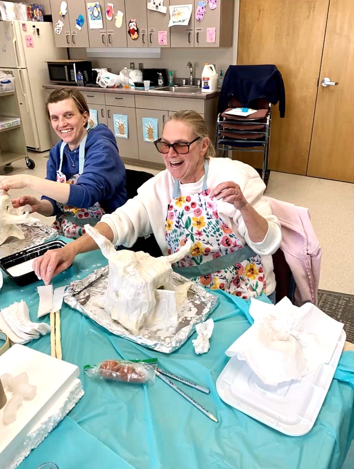 Two older women smile and laugh while creating gnome houses made from foil.